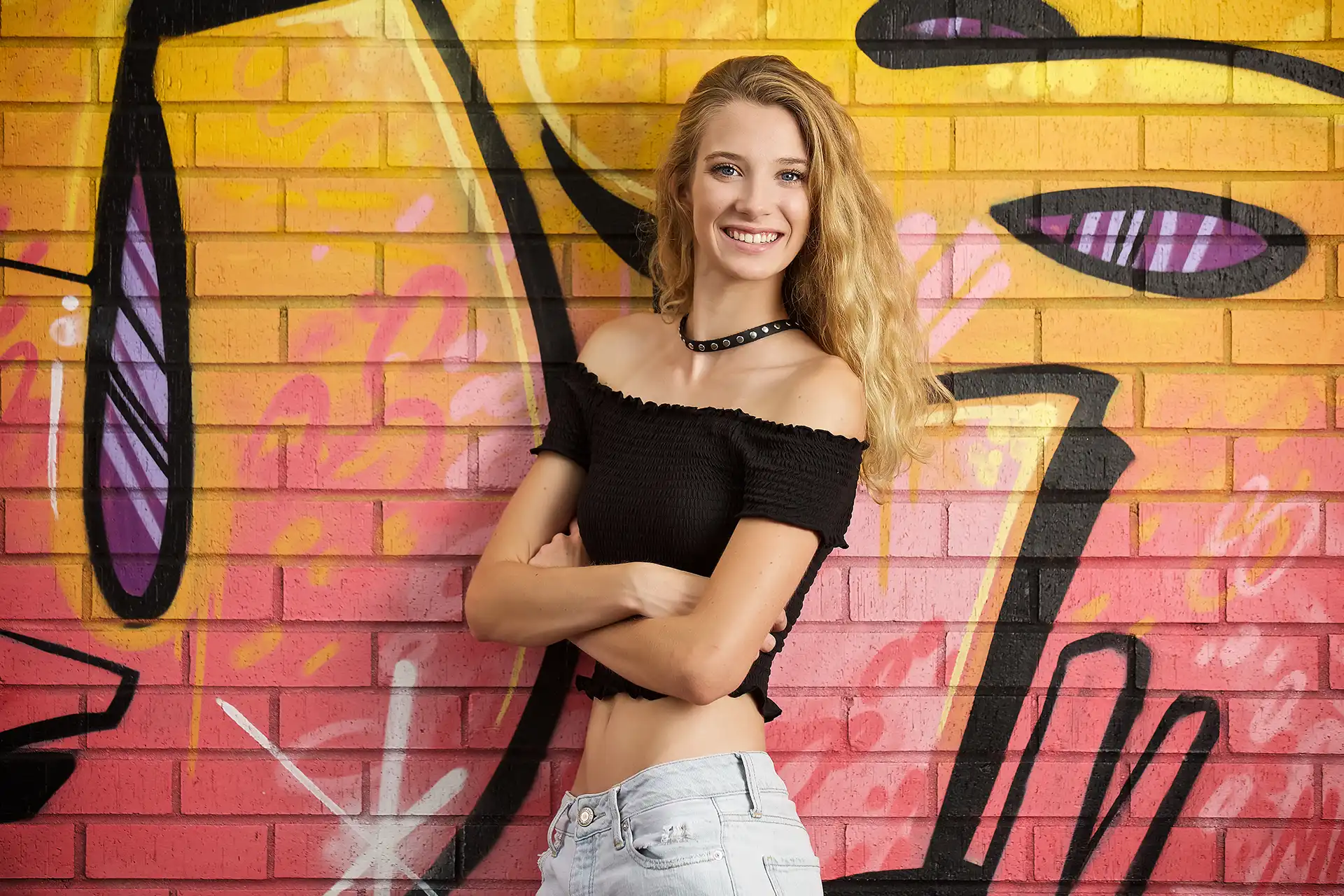 High school senior girl in black crop top and jeans in front of graffiti wall that reads Tulsa in yellow and pink paint. Tulsa photography