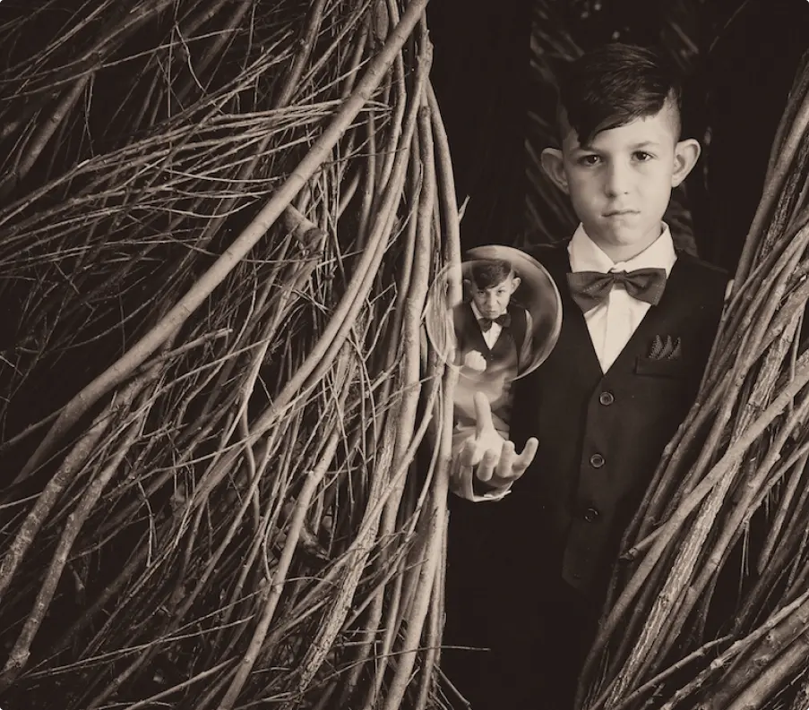 Sepia toned image of a boy in a vest and bow tie emerging from a hut made of woven sticks. He levitates a bubble that contains a small version of himself with an angry expression